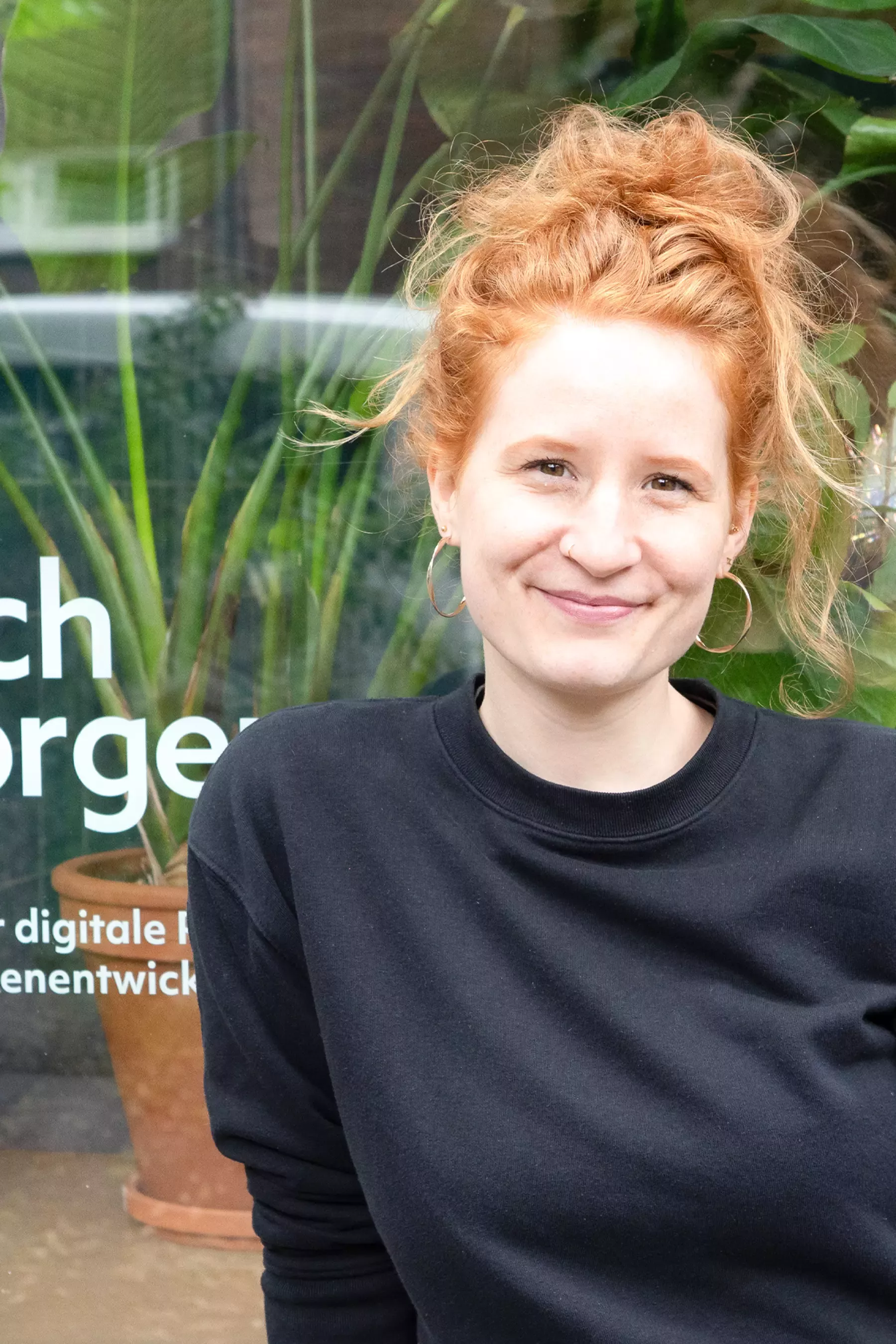 Selina is wearing a black sweater and standing in front of the office with a smile on her face. Behind her is a window with the words “nach morgen – Studio für digitale Produkt- und Marken­entwicklung” (nach morgen – Studio for digital product and brand development) written in white and a plant behind the glass