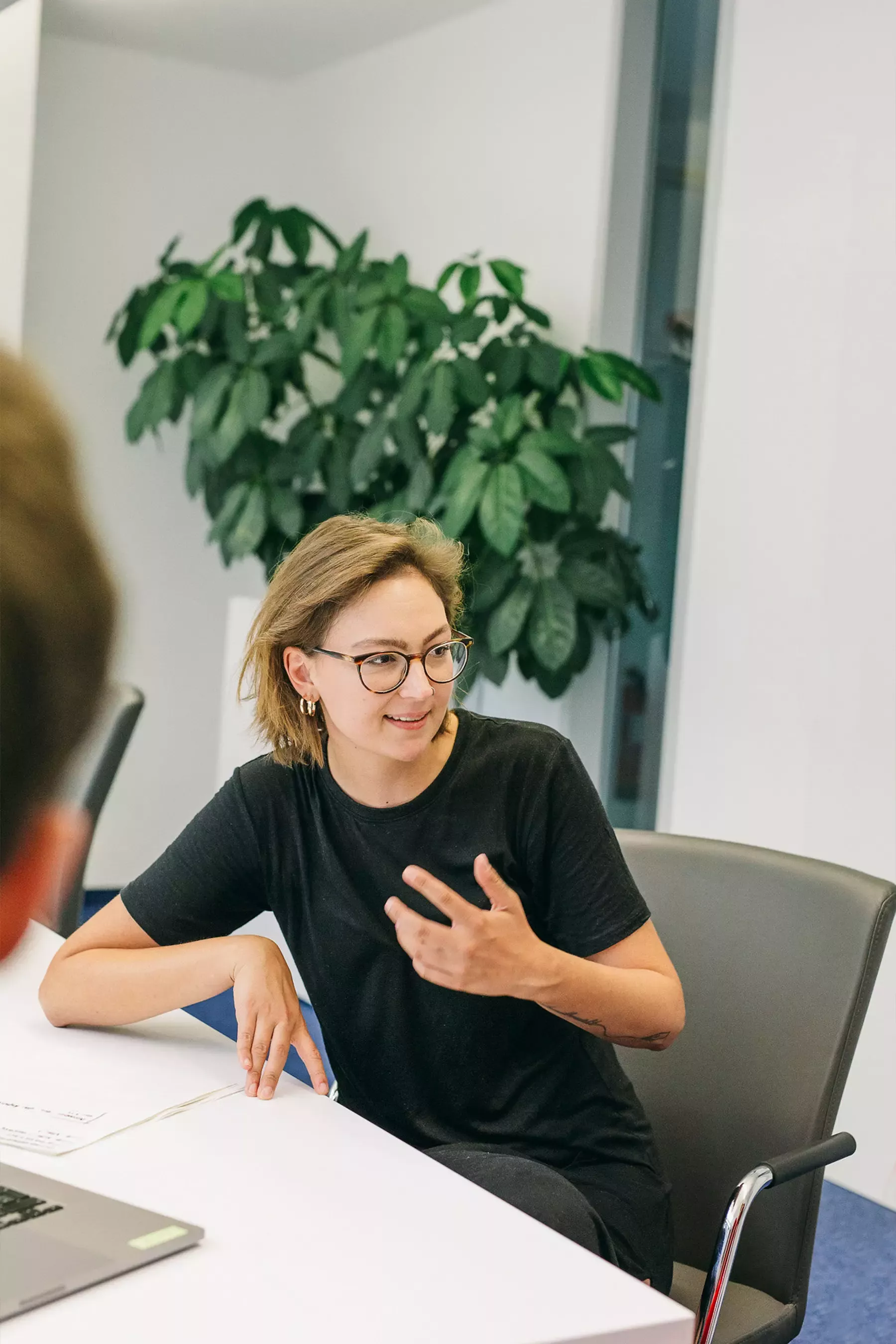 Lea sitzt an einem Tisch, gestikuliert beim Sprechen, im Hintergrund eine große grüne Pflanze