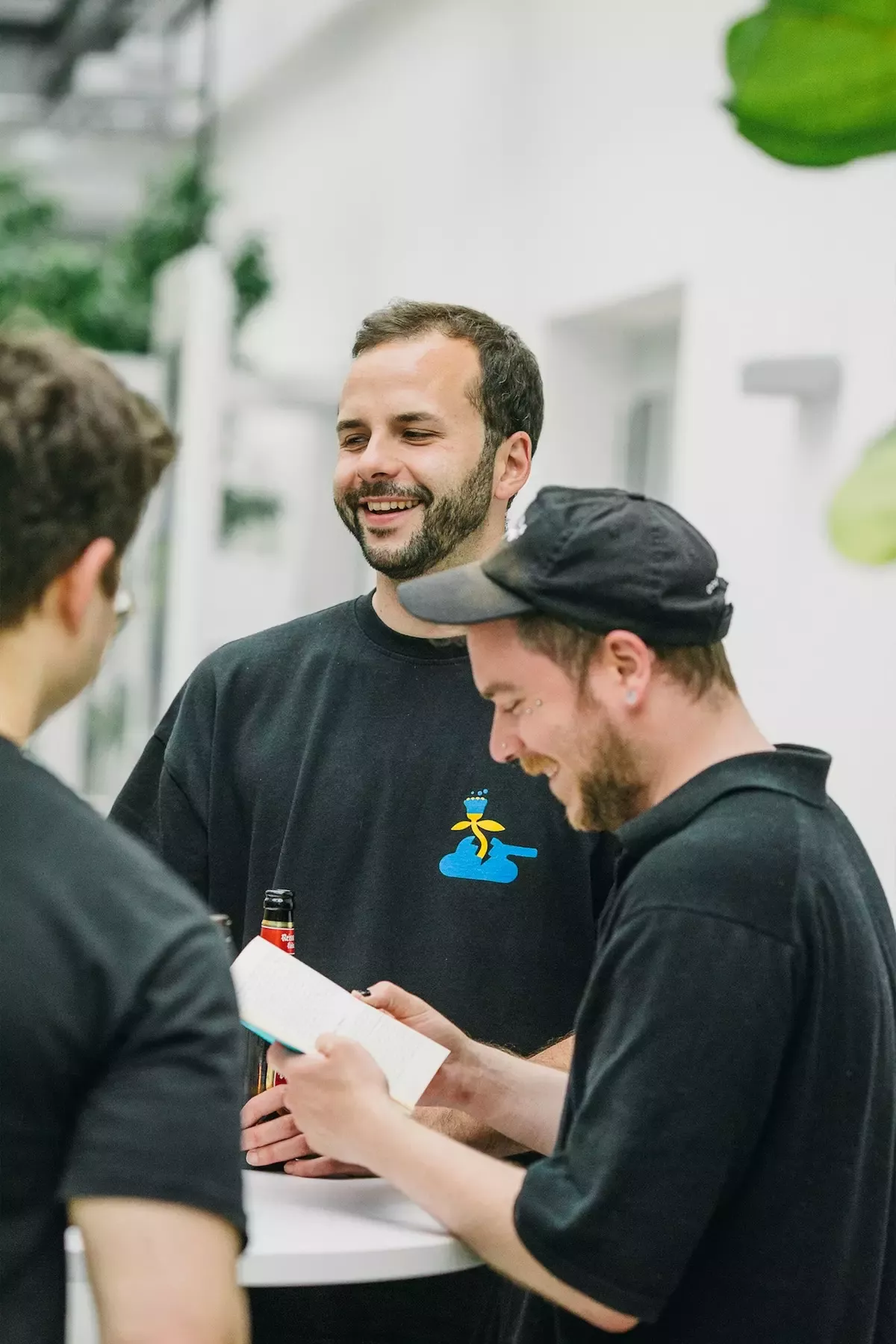 Nico, Steffen, and Tobi are standing together at a table, laughing and chatting. Tobi is holding a notebook, Steffen a drink