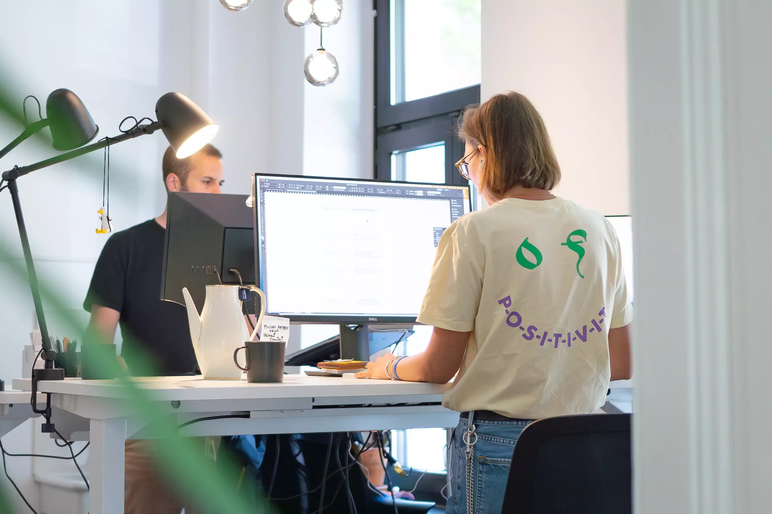 Lea and Steffen are working standing up at desks with monitors. Lea is in the foreground wearing glasses and a T-shirt with the words “P-O-S-I-T-I-V-I-T-Y” printed on it. Steffen is in the background wearing a black T-shirt.