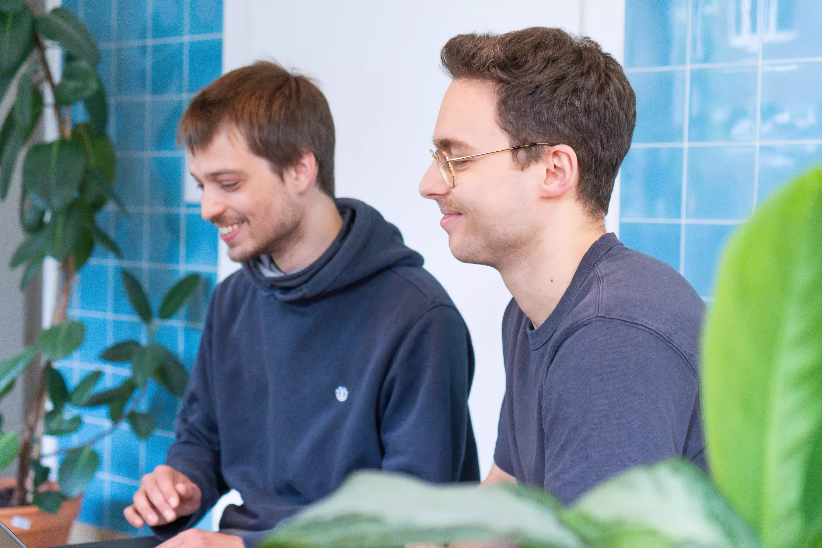 Jakob und Nico sitzen nebeneinander lachend vor einem Laptop in einem Raum mit blauen Kacheln im Hintergrund, Pflanzen im Vorder- und Hintergrund