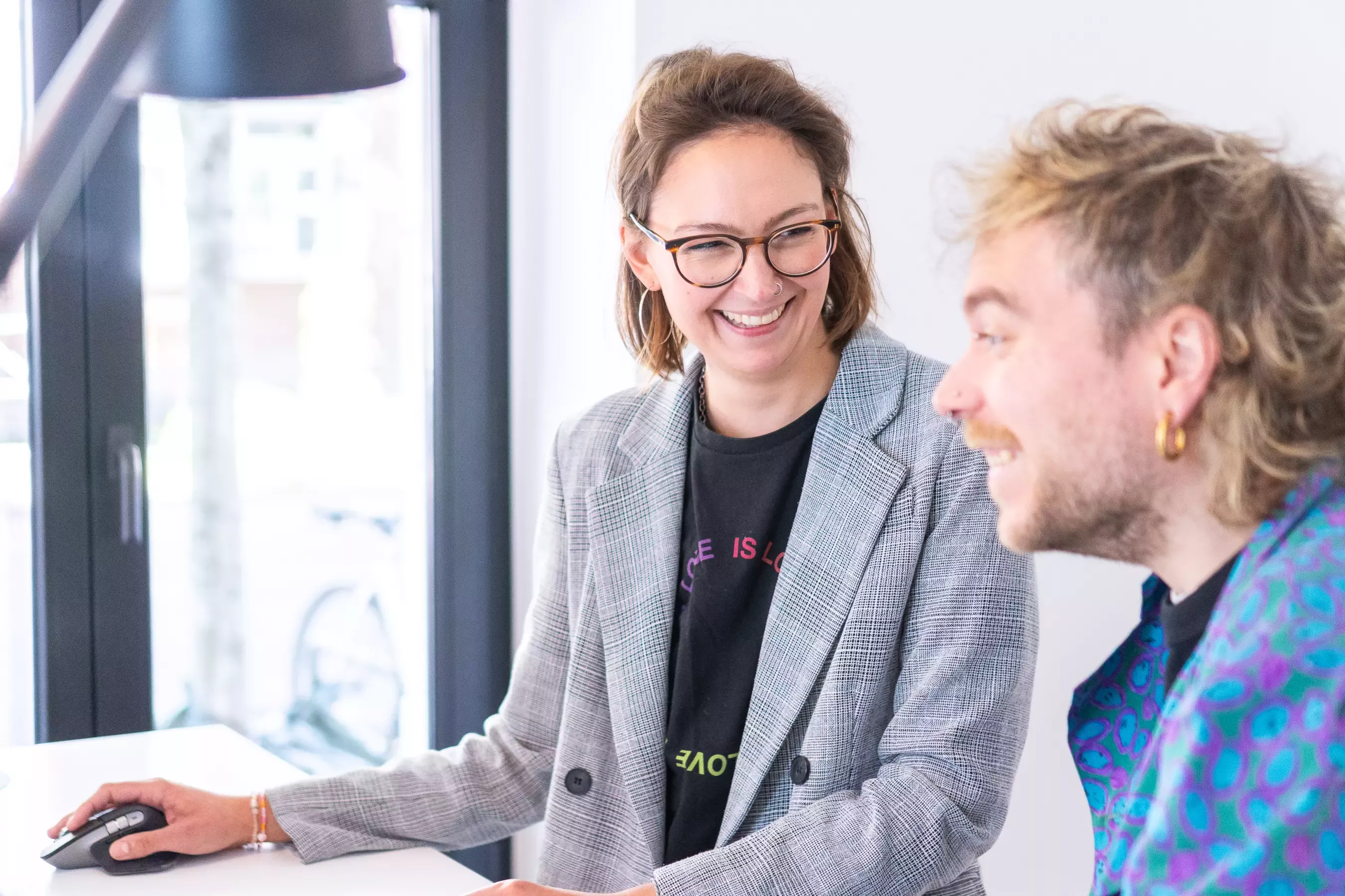 Lea and Tobi are standing next to each other at the desk, laughing. Lea is wearing glasses and a gray blazer, while Tobi is wearing a colorful patterned top