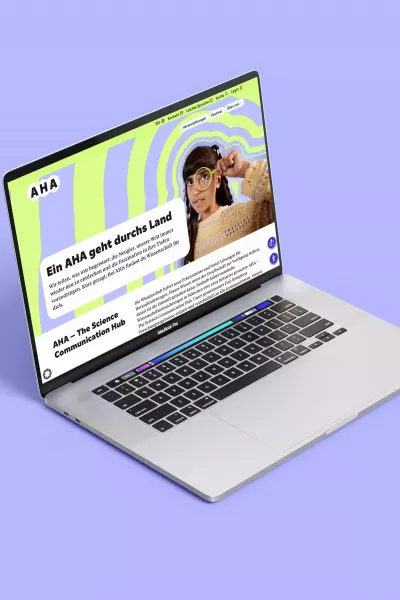 Geöffneter Laptop auf lilafarbenem Hintergrund, auf dem Bildschirm die AHA-Webseite mit grün-lila Muster, Person mit Lupe und Text \