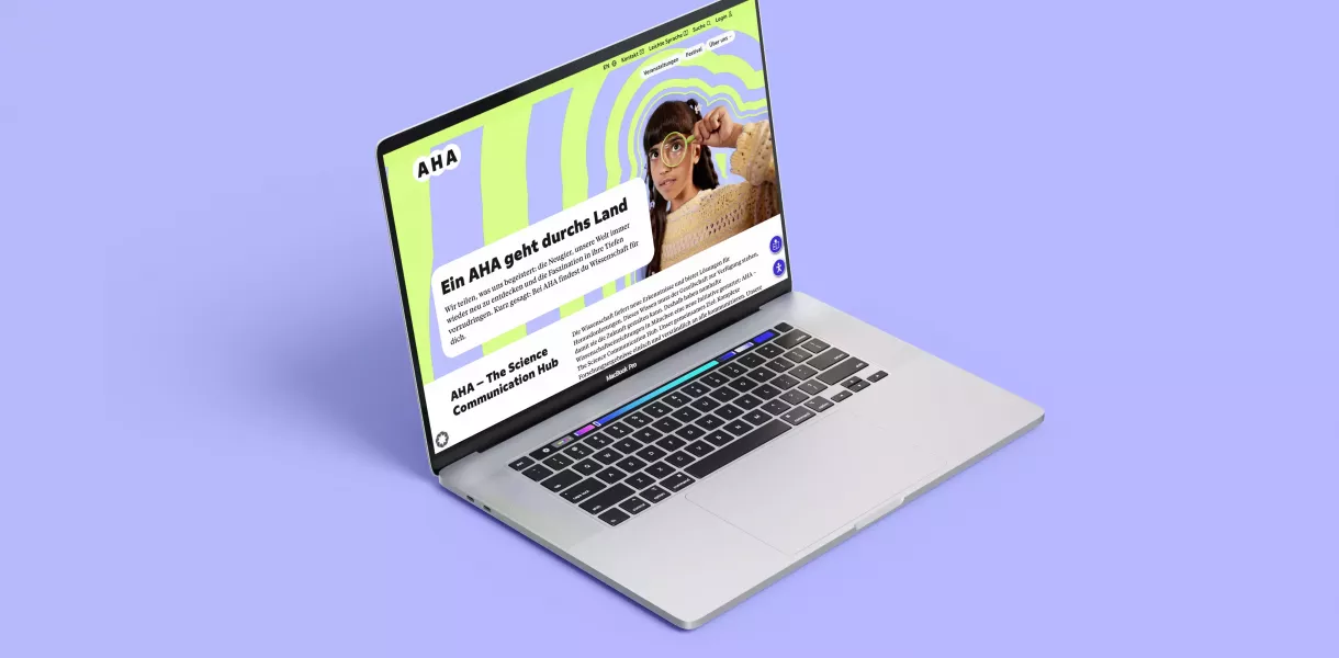 Geöffneter Laptop auf lilafarbenem Hintergrund, auf dem Bildschirm die AHA-Webseite mit grün-lila Muster, Person mit Lupe und Text \