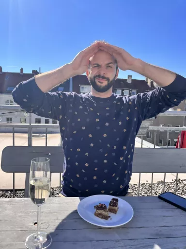 Steffen sitzt auf Dachterrasse des dimedis Bürogebäudes bei Sonnenschein, vor sich Glas Sekt und Teller mit zwei kleinen Kuchenstücken