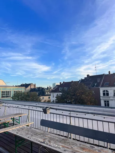 Blick von der Dachterrasse des dimedis Bürogebäudes mit Bierbänken und Lichterkette, darüber blauer Himmel und Wolken bei sonnigem Wetter