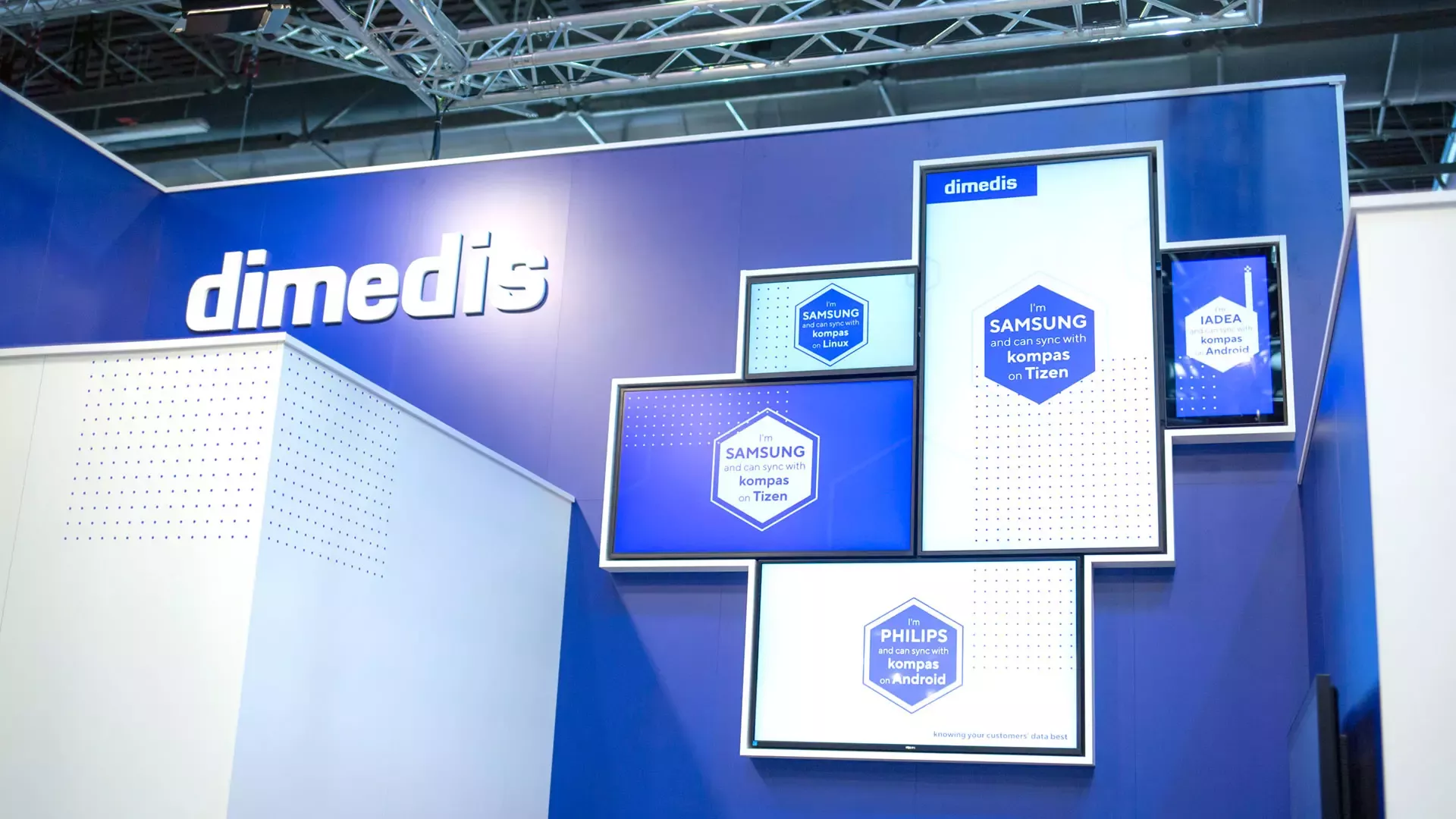Messestand von dimedis mit großem weißen Logo auf blauer Wand, daneben mehrere Bildschirme in verschiedenen Formaten mit blauen und weißen Hexagon-Grafiken und Texten zu Samsung, Philips und IADEA