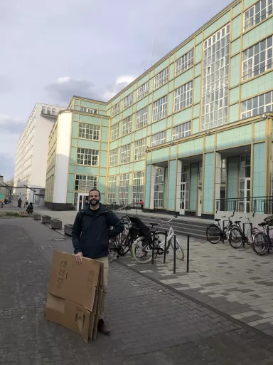 Steffen steht lächelnd vor dem dimedis Bürogebäude mit türkisfarbenen Fassadenplatten und vielen Fenstern, hält mehrere zusammengefaltete Kartons in der Hand
