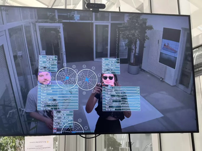 Monitor zeigt Live-Kameraaufnahme mit Tobi und Lea, deren Gesichter von Software erkannt und mit Daten zu Alter, Emotionen und Analyse-Grafiken überlagert werden