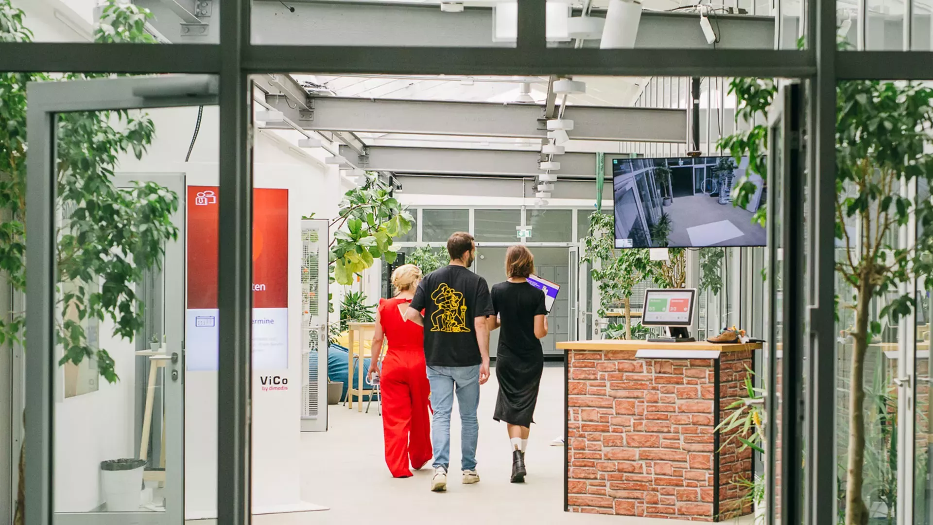 Das Foto zeigt den Eingangsbereich eines modernen, hellen Büros von dimedis. Lea und Steffen gehen mit einer weiteren Person durch den Flur. Rechts steht ein Tresen in Backsteinoptik mit einem Bildschirm, darüber hängt ein großer Monitor. Der Raum ist von vielen großen Pflanzen und Glasfronten geprägt
