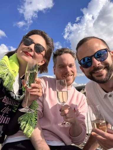 Lea, Tobi und Steffen sitzen draußen unter blauem Himmel mit Wolken, tragen Sonnenbrillen, halten Sektgläser und lächeln ind die Kamera