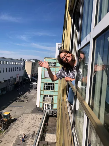 Lea lehnt sich aus einem Fenster des dimedis Bürogebäude und winkt lächelnd, darunter Blick auf eine Fußgängerzone mit Baustelle, Fahrrädern und Passanten bei sonnigem Wetter