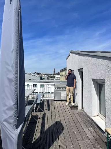 Dachterrasse mit Holzplanken, Bierzeltgarnitur und großem Grill, rechts Steffen am Handy, im Hintergrund Stadtdächer, eine Kirche und blauer Himmel