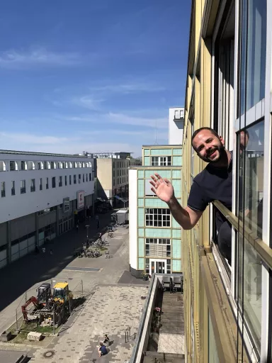 Steffen lehnt sich aus einem Fenster des dimedis Bürogebäude und winkt lächelnd, darunter Blick auf eine Fußgängerzone mit Baustelle, Fahrrädern und Passanten bei sonnigem Wetter
