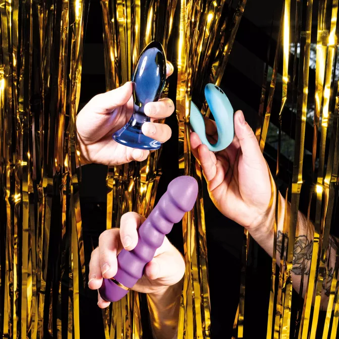 Three hands hold a purple dildo, a dark blue butt plug, and a light blue clitoral vibrator. Golden tinsel can be seen in the background