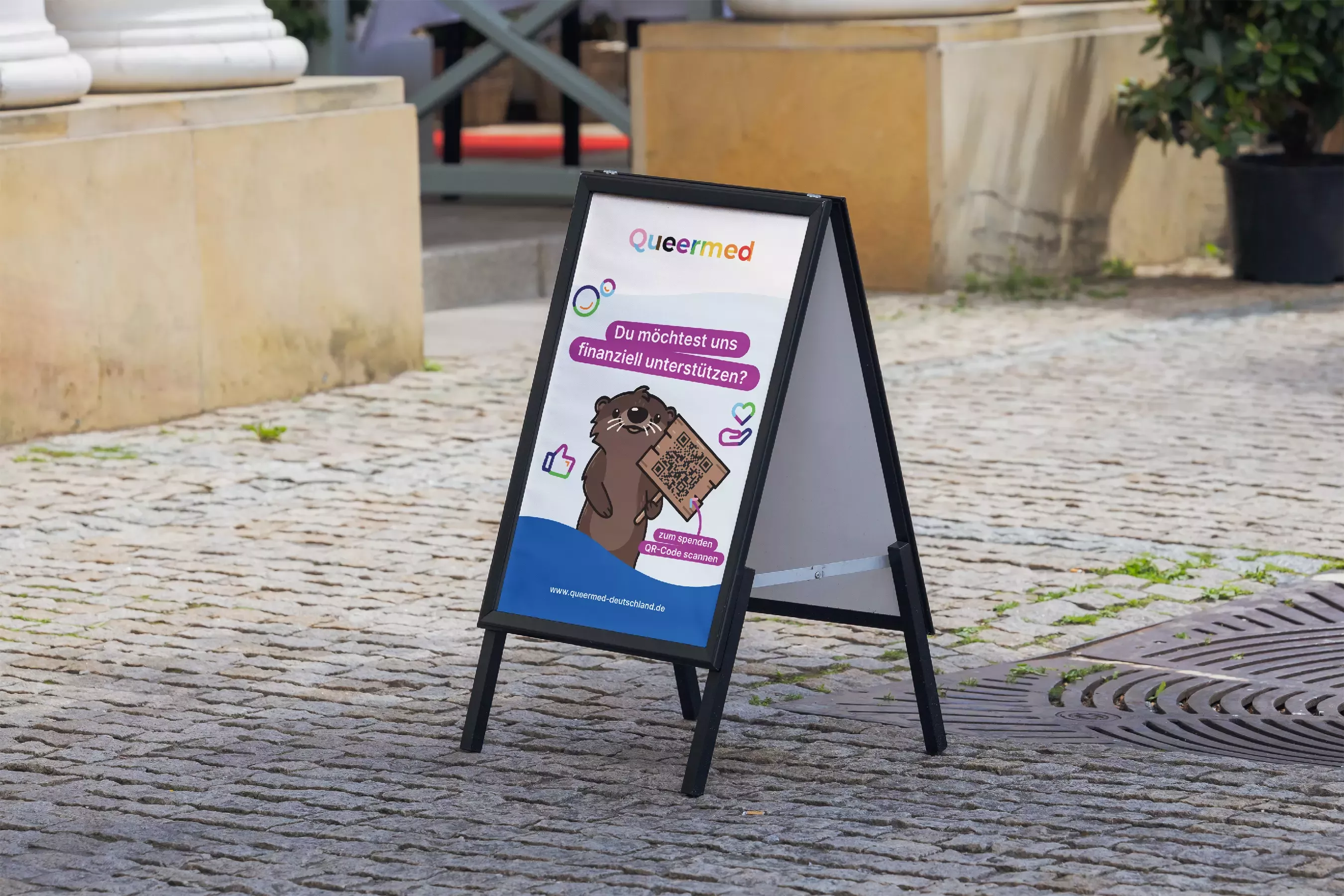 Display stand on cobblestones with Queermed logo, otter illustration, and text \