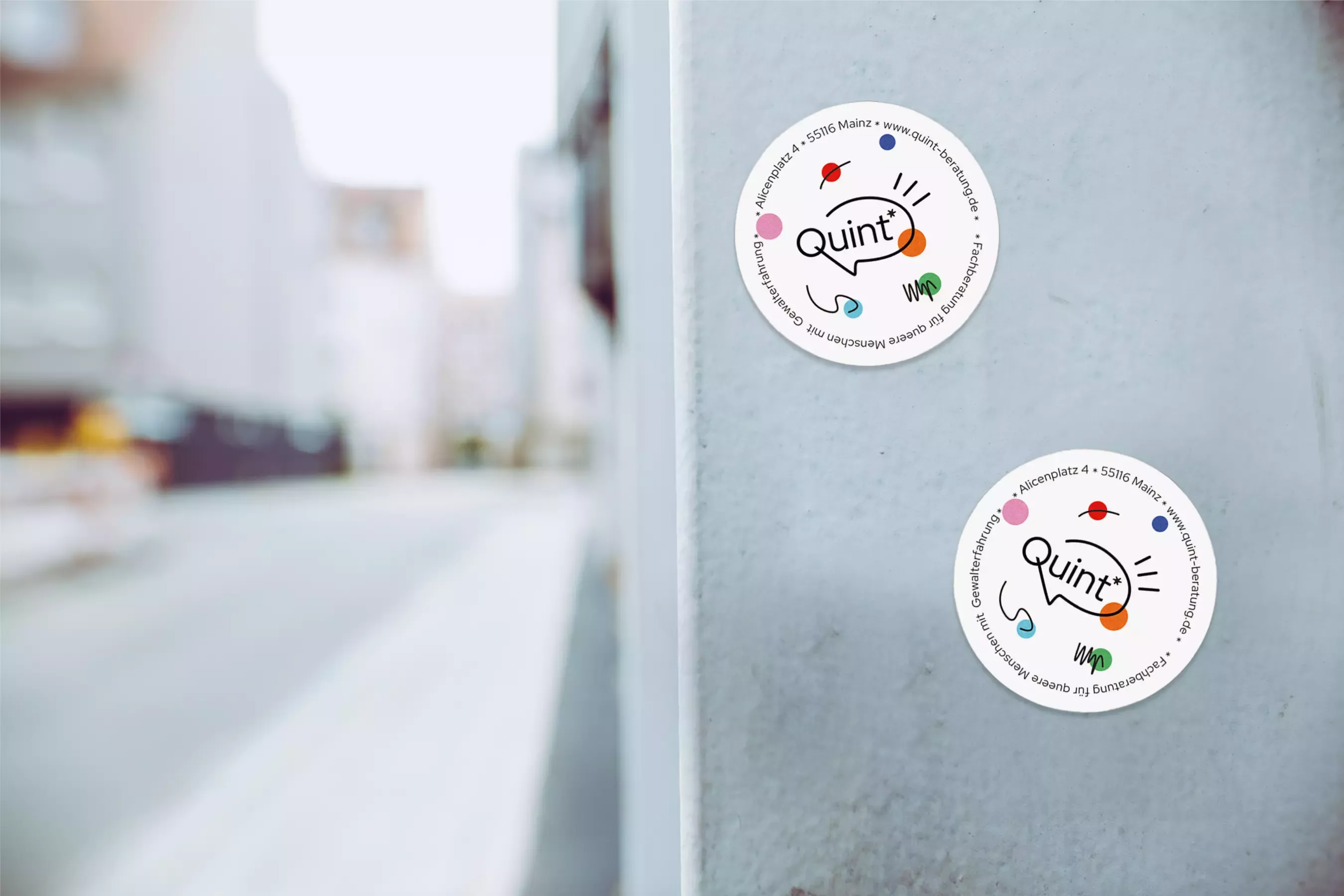 Round stickers with the Quint* logo, colorful line illustrations, and contact details, attached to a gray wall, with a blurred street with houses in the background