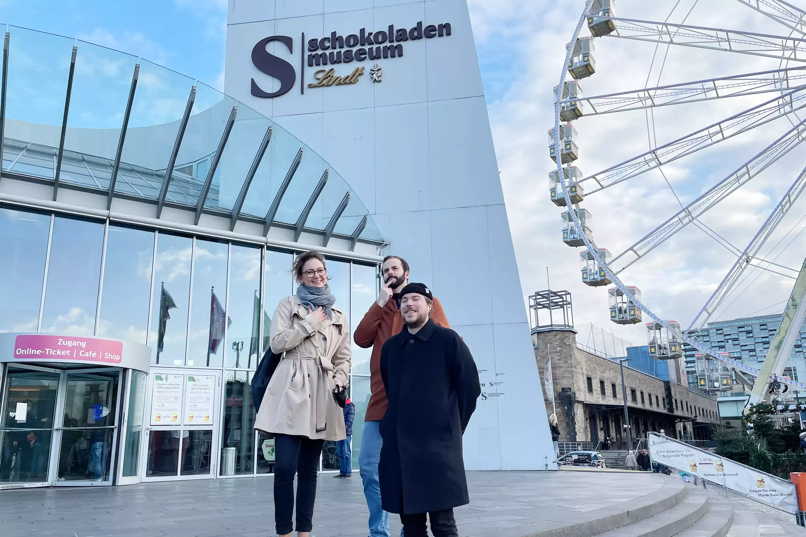 Lea, Steffen, and Tobi in front of the Chocolate Museum in Cologne. On the left is the entrance with a glass facade, next to it is the white museum building with the lettering \