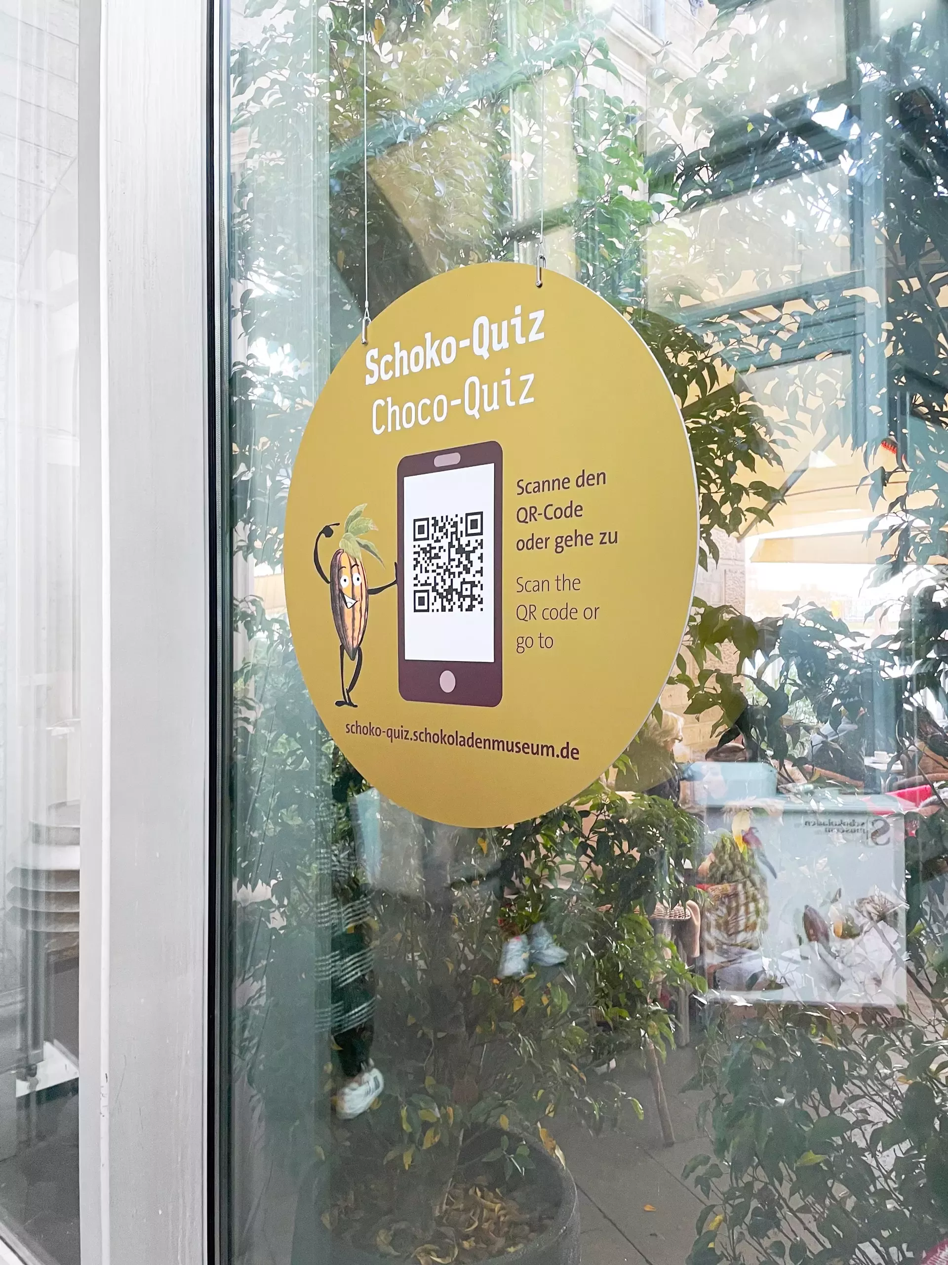Runder gelber Sticker auf Glastür mit der Aufschrift „Schoko-Quiz / Choco-Quiz“. Darunter eine Illustration einer Kakaobohne mit Gesicht, die auf ein Smartphone mit QR-Code auf dem Bildschirm zeigt