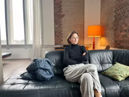 Person sitzt entspannt auf schwarzem Ledersofa mit verschränkten Armen, neben sich Jacke und Kissen, im Hintergrund rote Ziegelwand große Fenster die den Raum erhellen