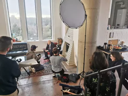 Videoproduktion in hellem Loft mit großen Fenstern, mehrere Personen mit Kamera, Licht und Reflektoren, eine Person sitzt im roten Stuhl vor Kamin und liest etwas vor