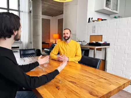 Steffen und eine Person im Gespräch an großem Holztisch, Hintergrund heller Raum mit hohen Fenstern und Betonpfeilern