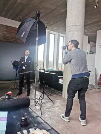 Fotoproduktion im Loft, Fotograf mit Kamera richtet sich auf eine Person im Anzug aus, die vor grauem Hintergrund und Softbox steht und lächelt. Im Hintergrund große Fenster und Betonsäule