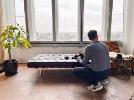 Person kniet vor einer schwarzen Ledercouch mit Kamera in der Hand, ruhige Atmosphäre und hell durch große Fenster im Hintergrund, Pflanze links, Stuhl und Tisch rechts