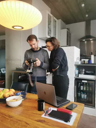 Lea und eine weitere Person stehen am Holztisch in Küche, beide schauen gemeinsam auf eine Kamera, auf dem Tisch Laptop, Obstschale und Notizbuch
