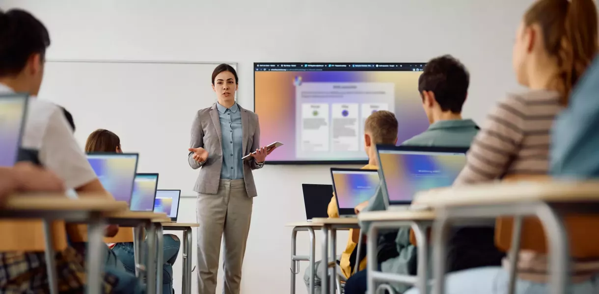 Teacher stands in front of a class with a tablet, presenting content on a large screen; several students sit at tables with laptops