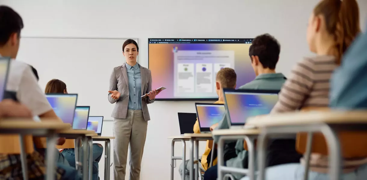 Lehrer*in steht mit Tablet vor einer Klasse, präsentiert Inhalte auf großem Bildschirm, mehrere Schüler*innen sitzen mit Laptops an Tischen