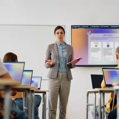 Teacher stands in front of a class with a tablet, presenting content on a large screen; several students sit at tables with laptops