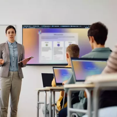 Lehrer*in steht mit Tablet vor einer Klasse, präsentiert Inhalte auf großem Bildschirm, mehrere Schüler*innen sitzen mit Laptops an Tischen