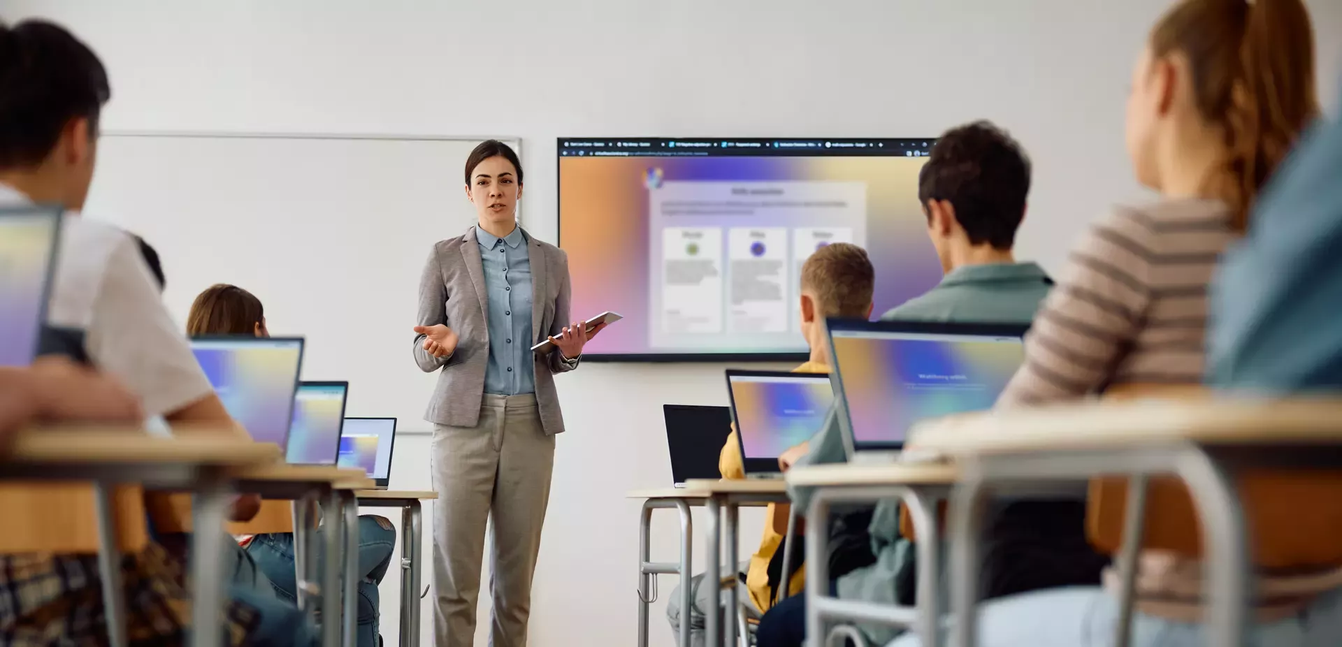 Teacher stands in front of a class with a tablet, presenting content on a large screen; several students sit at tables with laptops