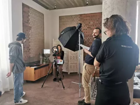 Mehrere Personen in einem hellen Raum bei einer Videoproduktion mit Kamera, Stativ, Ringlicht und Softbox, eine Person sitzt vor der Kamera. Steffen und Lea stehen im Vordergrund während Steffen ein Skript in Händen hält und in die Kamera lächelt. Im Hintergrund rote Backsteinwand und große Fenster