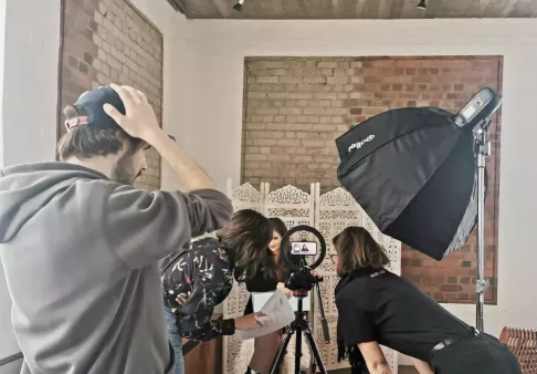 Videodreh mit Kamera auf Stativ, Ringlicht und Softbox, mehrere Personen beugen sich über Skript, im Hintergrund sitzt eine Person vor Paravent und spricht in die Kamera