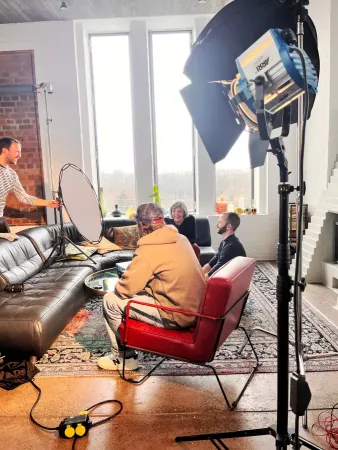 Mehrere Personen sitzen in einem gemütlichen Loft auf Sofa und Stühlen, Filmlicht und Reflektor aufgebaut, große Fenster und rote Backsteinwand im Hintergrund