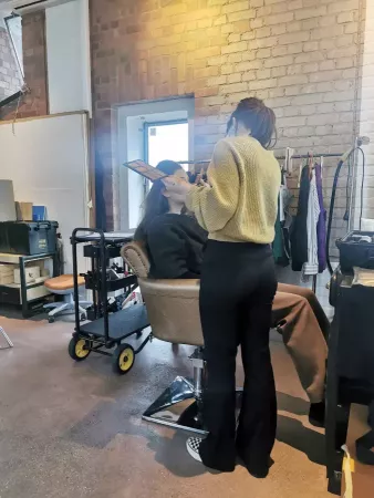 Eine Person sitzt auf einem Stuhl, während eine andere Person mit Pinsel und Palette Make-up aufträgt, im Hintergrund Kleiderstange und Backsteinwand