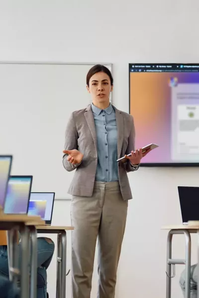 Teacher stands in front of a class with a tablet, presenting content on a large screen; several students sit at tables with laptops