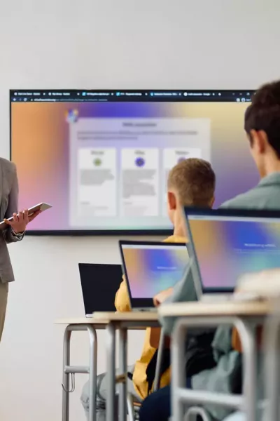 Lehrer*in steht mit Tablet vor einer Klasse, präsentiert Inhalte auf großem Bildschirm, mehrere Schüler*innen sitzen mit Laptops an Tischen