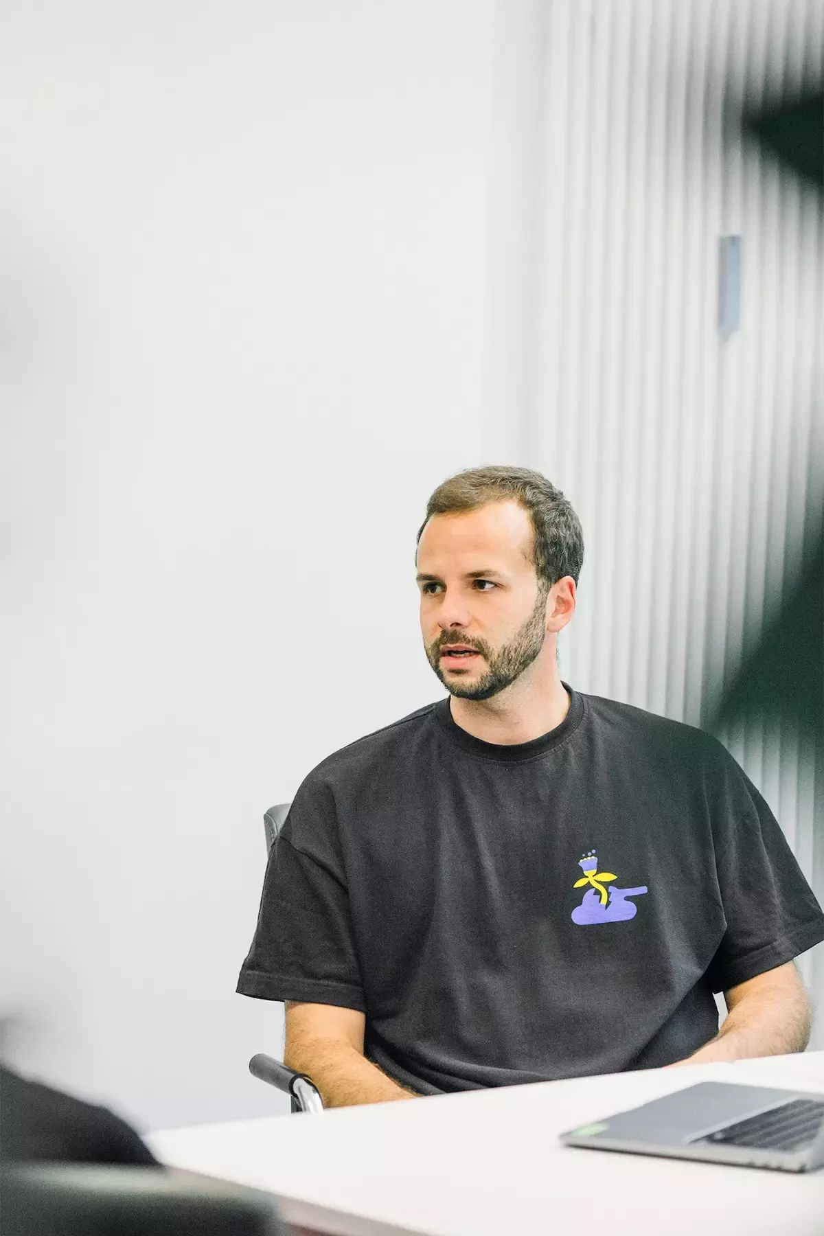Steffen is sitting in the meeting room in front of a white wall, wearing a black T-shirt with a yellow and purple motif, with an open laptop in front of him