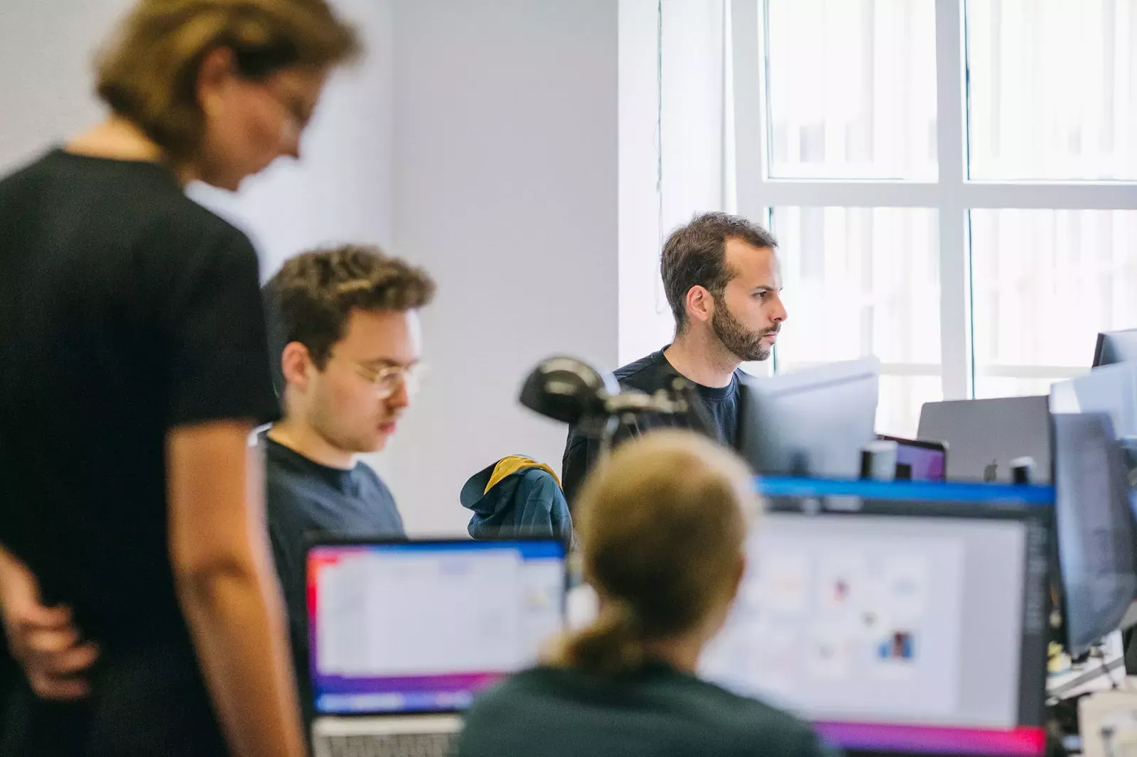 Several people are working intently at their desks with monitors in a bright office