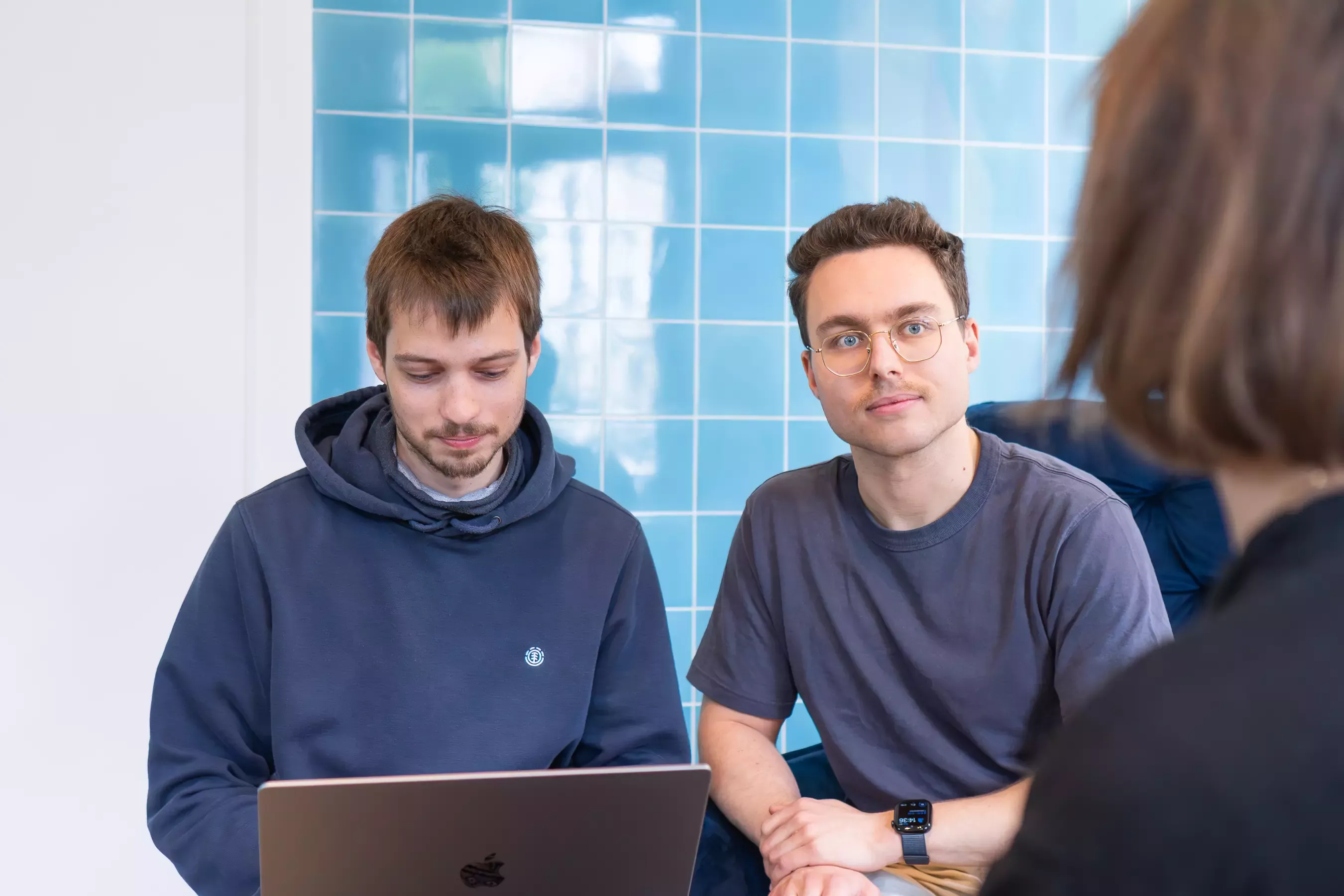 Jakob, Nico, and Lea are sitting in front of a blue tiled wall. Jakob is busy with a laptop, while Nico and Lea are talking