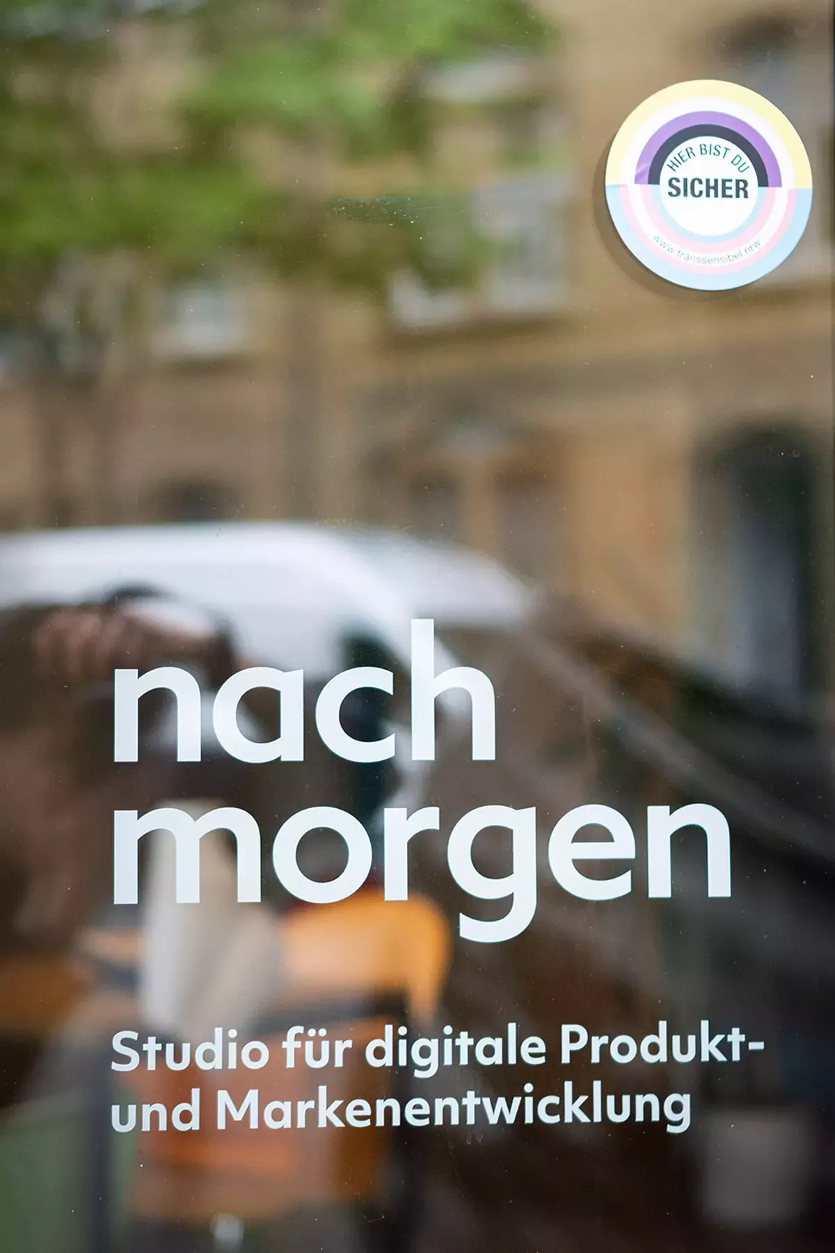 Lettering “nach morgen – Studio für digitale Produkt- und Markenentwicklung” (nach morgen – Studio for digital product and brand development) on glass front, next to it sticker “Hier bist du sicher” (You are safe here) in purple, white, and blue colors representing the trans flag