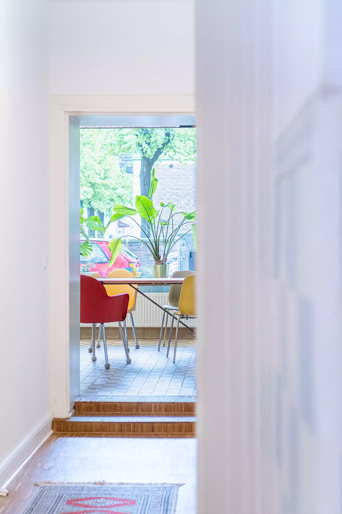 View through a hallway into a bright room with colorful chairs, plants, and a window front overlooking trees and a parked car