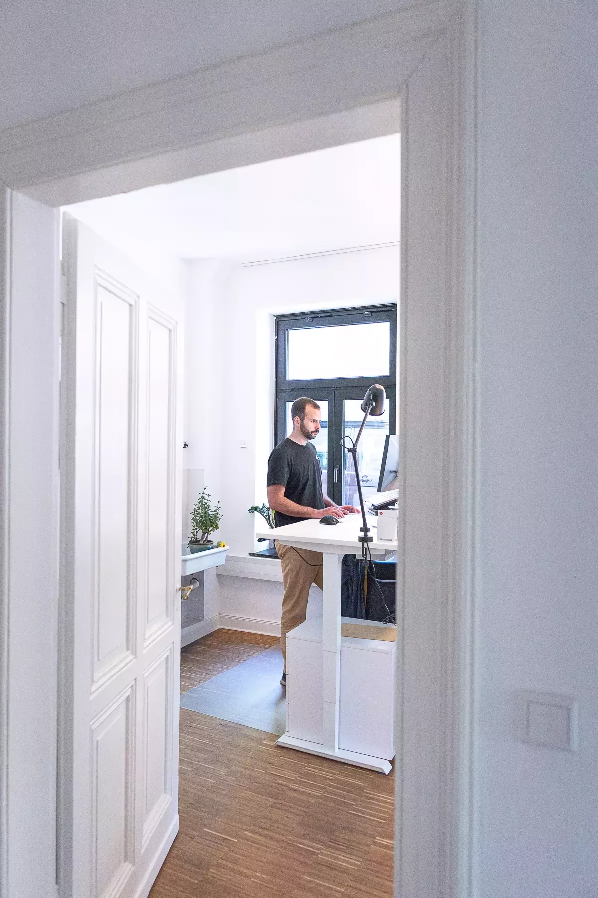 Steffen works intently while standing at a height-adjustable standing desk in a bright office with plants and a large window, photo taken through a door frame