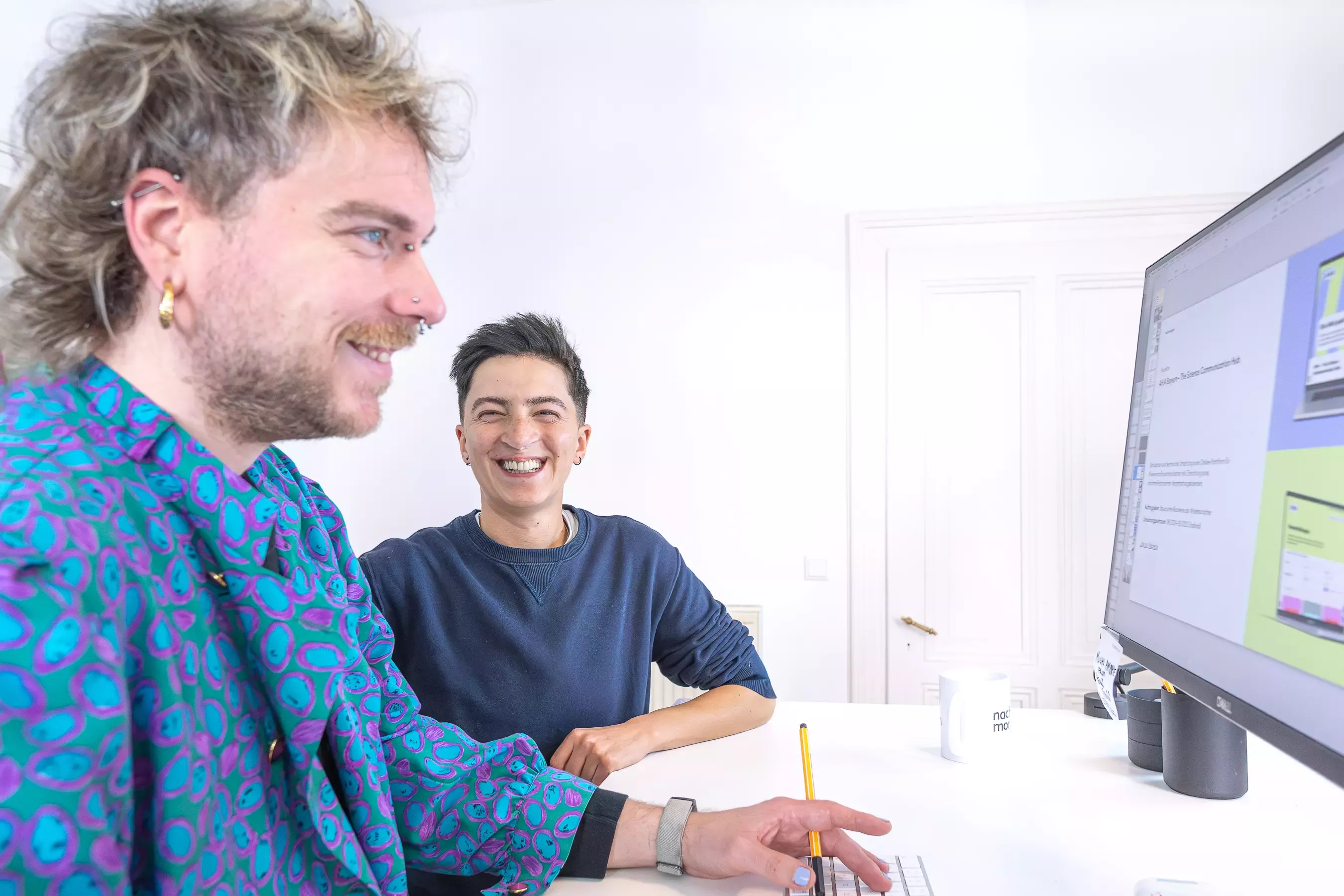 Tobi and Yase sit next to each other at the desk, laughing, and look together at a large monitor