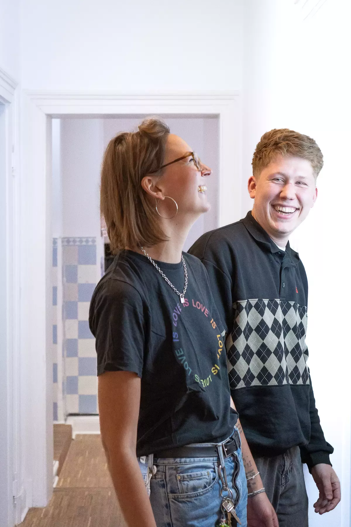 Lea and Maxi walk down the hallway laughing. Lea wears glasses and a black T-shirt with “Love is” printed on it, Maxi wears a black and gray sweatshirt