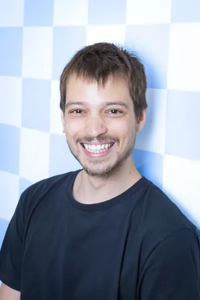 Portrait of Jakob with short dark hair and beard wearing a black T-shirt, smiling in front of a blue and white checkered background in a bright setting