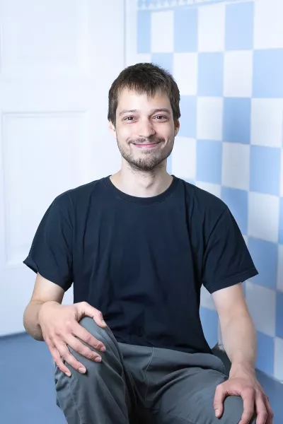 Jakob sits in a relaxed position, smiling in the agency in front of a blue and white checkered background, with short dark hair and a beard, wearing a black T-shirt